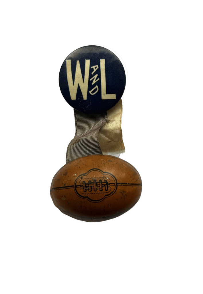 A small, vintage Washington and Lee football commemorative pin displayed vertically. The top is a round, navy blue button with the letters "W L" and "AND" in small text written diagonally between them in a cream color. A piece of fabric ribbon connects the button to a small, brown, three-dimensional football charm at the bottom.