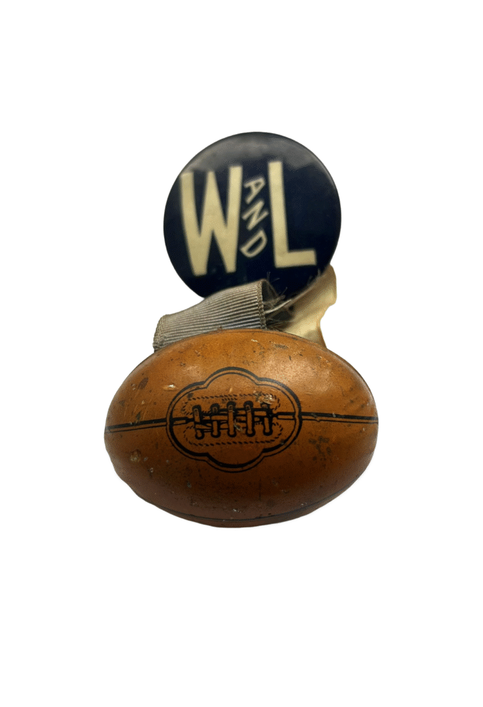 A close-up view of a small, vintage Washington and Lee football commemorative pin. The top is a round, dark blue button displaying the large cream letters "W" and "L" with the word "AND" curved between them. A frayed ribbon attaches the button to a small, brown, football-shaped charm with painted laces.