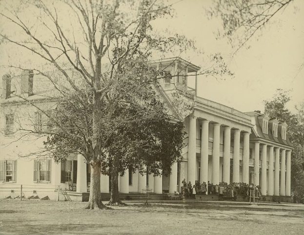 A sepia-toned photograph of a stately, multi-story building featuring a prominent white colonnade, two-story portico, and dormer windows. A large group of students or people is gathered on the steps, with large, bare trees framing the foreground.