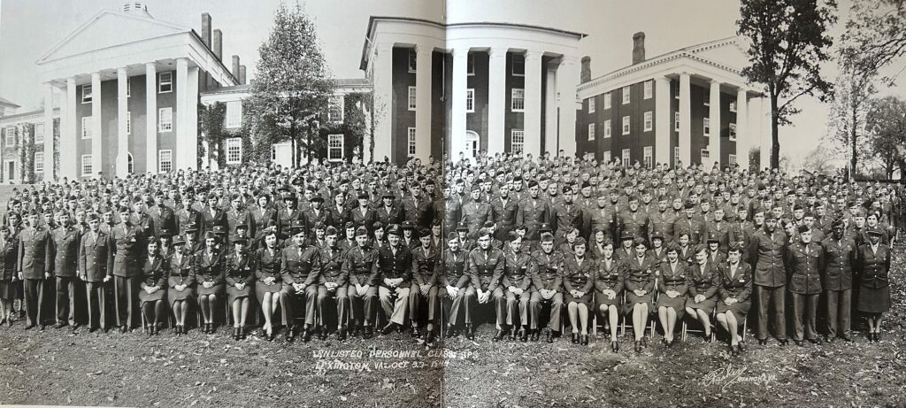 A wide, panoramic black and white photo of a large group of over two hundred men and women, all in military uniforms, posing outside on a lawn. The backdrop features three large, classical-style university buildings with prominent white columns. Text written below the group identifies them as the "WLU Enlisted Personnel Class S-8, Lexington, VA, Oct 25–Dec 18, 1943."