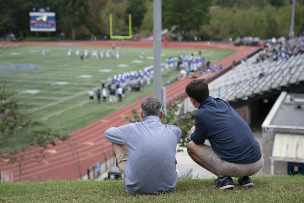 A contemporary photograph taken from a high vantage point overlooking the W&L football stadium and field. In the foreground, a man and a younger person are seated on the grass, seen from behind, watching the game in the distance. The background shows the field with players in blue and white uniforms, a red track, and a scoreboard on the far left, suggesting a game during Family Weekend.
