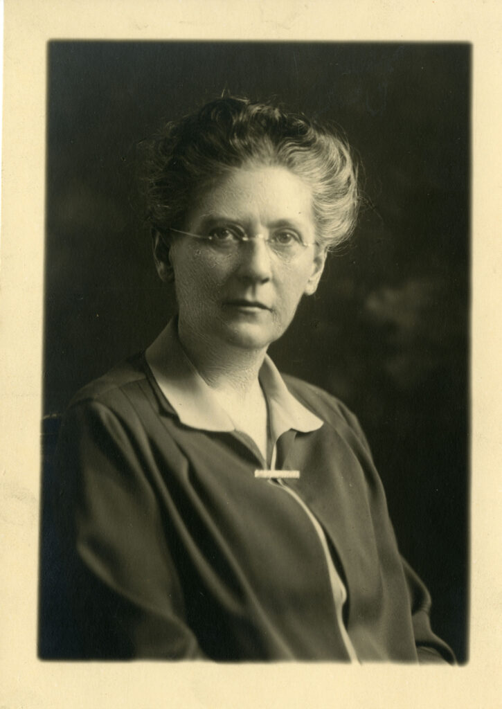A black and white studio portrait of Lucy Ella Fay. She is a middle-aged woman with her hair pulled back and wearing round spectacles. She is facing the camera, wearing a dark jacket or cardigan over a light-colored collared shirt, fastened at the neckline with a small, light-colored brooch or clip. The background is dark and uniform.