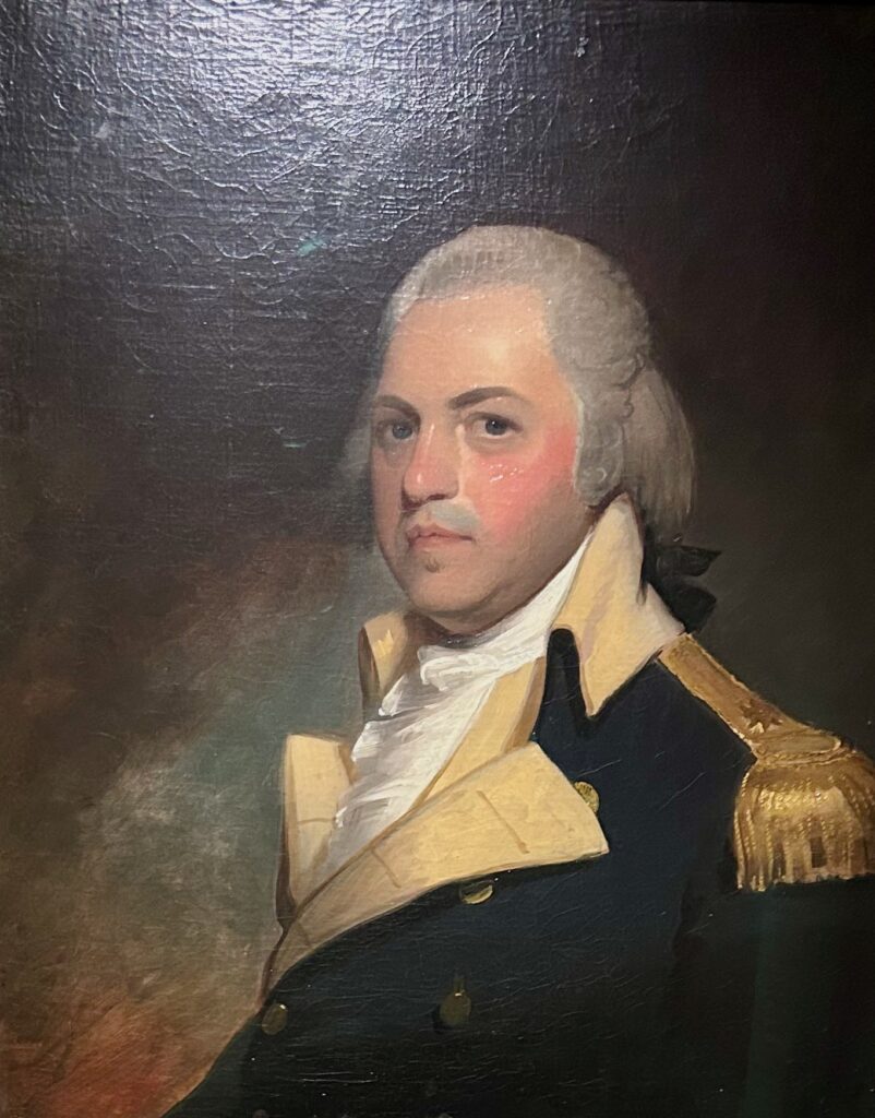 Portrait of Henry "Light-Horse Harry" Lee in his Continental Army uniform. He is a white man wearing a white powdered wig, with red cheeks, and a dark jacket. There is gold detail on his shoulders and he his wearing a white collared shirt.